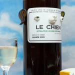 Installation featuring two bottles of wine with dog faces, two sandwiches with dog faces, a picnic basket with a baguette, an apple and a pear all with dog faces on top of a red and white checkered table clothe and a painted sky with clouds in the shape of dogs.