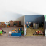 miniature diorama by Abigail Goldman of the interior of house with three rooms, one in which a family is eating dinner and another with people chained and bleeding