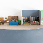miniature diorama by Abigail Goldman of the interior of house with three rooms, one in which a family is eating dinner and another with people chained and bleeding