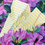 a paper airplane made of yellow lined paper with a doodle on it resting in a bed of purple flowers