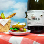 Installation featuring two bottles of wine with dog faces, two sandwiches with dog faces, a picnic basket with a baguette, an apple and a pear all with dog faces on top of a red and white checkered table clothe and a painted sky with clouds in the shape of dogs.