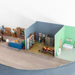 miniature diorama by Abigail Goldman of the interior of house with three rooms, one in which a family is eating dinner and another with people chained and bleeding