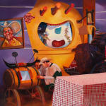 Painting of a television set with three men dressed as cowboys standing around a table with a red and white checkered table cloth. In the background is a painting on the wall of a cowboy and other western paraphernalia