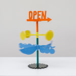 Metal sculpture with the word OPEN on the top and an arrow in orange. Stacked below are four yellow circles on yellow rods and stacked below those are blue berries and bananas on a steal stand.