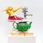 green teapot with a dog face spitting up tea and shoes, a hair dyer, a faucet spout and a baseball stacked on top