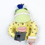 Ceramic creature with large lips holding a book and a Dunkin Donuts bag and wearing a green visor