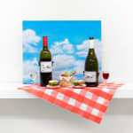 Installation featuring two bottles of wine with dog faces, two sandwiches with dog faces, a picnic basket with a baguette, an apple and a pear all with dog faces on top of a red and white checkered table clothe and a painted sky with clouds in the shape of dogs.