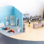 miniature diorama by Abigail Goldman depicting three scenes, two people dead in a bathroom, a laboratory and a family watching tv