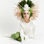 Photo of a woman in green tights and a white dress wearing a bonnet with leaves and sticks on it