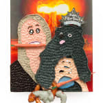 Mixed media hanging sculpture of a woman with a carrot in her mouth holding a small black dog with a Happy New Year paper crown on his head and a pretzel and a carrot in his hands. In the background an atomic bomb is going off.