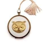 gold round cosmetic case with white middle and a gold owl in the center and a powder pink tassel attached the side