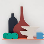 Sculpture of an orange shelf holding a pile of three limes, a black vase, a brown vase, two white plates stacked on top of each other and a blue mug.