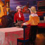 Painting of a television set with three men dressed as cowboys standing around a table with a red and white checkered table cloth. In the background is a painting on the wall of a cowboy and other western paraphernalia