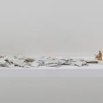 Digital photograph of an installation piece in which multiple pieces of broken ceramic shards are arranged in a horizontal pile. The ceramic pieces are placed on top of a white wooden shelf which is mounted onto a white wall. Towards the right of the piece a small metallic golden figurine with a watering can stands above some of the ceramic shards