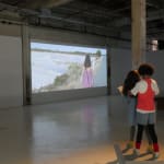 Still from video playing in gallery; a girl walks along a road.