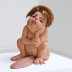 Sculpture of squatting anthropomorphic creature with human skin, webbed feet, rodent-like teeth and voluminous hair. Creature rests its head on its hand and gazes into the distance. From its groin area emerges an oversized, hairy tongue.