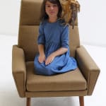 Sculpture of girl in blue dress seated on green chair in kneeled position with hands in its lap. On girl’s left shoulder is sculpture of owl-like creature with one wing outstretched to protect the girl’s head.