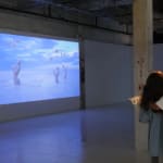 Still from video playing in gallery; a girl in a pink dress stands in a sand flat, watching bubbles blossom from three white, alien plants.