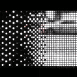 Gif of rectangular piece with multiple LEDs behind Plexiglass that displays a highly pixelated and blurred city street scene The people to the right side of the image are black and white and have more clarity while the figures at the left side are colorful but very blurred and abstracted