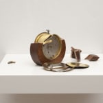 Digital photograph of an installation piece of a thick white wooden shelf which holds a broken and dismantled round wooden and metal clock. On top of the clock is a tiny human figurine who is crouched down with a tool in hand