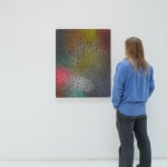 A man stands to the right and faces a multicolored, brightly colored abstract painting, which hangs on a white wall