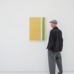 A man stands to the right and faces a yellow, white, green and grey abstract painting mimicking fabric.