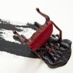 Detail of one of the several figurines. This one is of a toppled over brown chair. Underneath the chair is scattered black paint