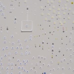 Detail; scattered square white cutouts with blue, red, and yellow dots on a white background with a 3D white flap left of center.