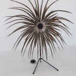 Electronic sculpture installation which is placed in an open white walled gallery space. The sculpture consists of a tall central metal pole which spreads out into multiple feathers at its top. The sculpture mimics the forms of a full bloom flower
