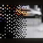 Still image of a rectangular piece with multiple LEDs behind Plexiglass that displays a pixelated street scene To the right side the scene is in black and white and has more clarity due to a higher density of LEDs while the scene at the left is colorful but very pixelated due to fewer LEDs