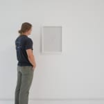 A man stands to the left of a framed white vertical rectangle hanging on a white wall.
