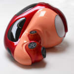 Sculpture of shiny orange and red automotive figure with four wheels lying in fetal position.
