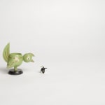Still from video; green bird figurine bends towards small penguin figurine with outstretched wing on white backdrop
