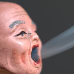 Still image from the video Actualidades/Breaking News. In the still a wrinkled gray haired human figurine head is breathing in or expelling a stream of smoke