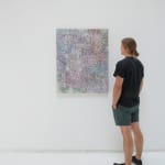 A man stands to the right and faces a multicolored, brightly colored abstract painting that hangs on a white wall.