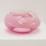 UFO-shaped glass sculpture. Top 2/3 opaque blush-pink, bottom 1/3 translucent pink. Clear top with clear bubbles of varying sizes, on white pedestal.