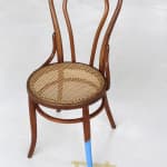 Digital photograph of an installation piece in which one half of a regular sized wooden chair is painted a bright baby blue. An incredibly tiny human figurine with a paint roller is placed next to the blue leg and is assumed to be the one responsible for painting it. Surrounding the painted leg and the tiny figurine is an uneven square of beige painters tape
