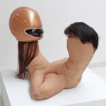 Sculpture of two conjoined thumb-shaped bodies made of human skin. One has long hair and wears a shiny orange helmet, and the other has short wavy black hair. Though they do not have features, the two shapes appear to be looking at each other and are conjoined in by their groin areas.