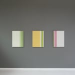 Three abstract paintings mimicking fabrics hang side by side on a grey wall.