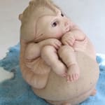 Sculpture of small fetus-like creature with human skin, hair, hands and feet. Creature has rodent face and is encased in a rounded spiked shell. Creature sits on fuzzy blue carpet.