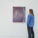 A man stands to the right and faces a multicolored abstract painting, which hangs on a white wall.