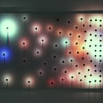 Still image of a rectangular aluminum board with metal rods arranged more densely towards the right side. On the ends of each rod multi-colored LEDs are mounted and they shine soft glowing circles on the surface of the board