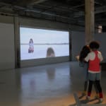 Still from video playing in gallery; a girl stands in a sand flat, looking at a small, hairy animal.