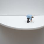 Digital photograph of an installation piece in which small human figurine bends over to pick up a tiny silver metallic bulb off of the surface of a white semi circle wooden shelf