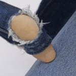 Detail of what looks like a finger whose knuckle looks like a knee protruding from hole in denim.