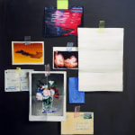 Blurred items taped to dark gray wall, including: picture of a vase of flowers, other abstracted images, postcards, a letter, and other pieces of paper.