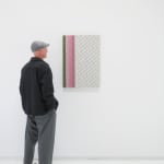 A man stands to the left and faces a green, pink, and white abstract painting mimicking fabric, which hangs on a white wall.
