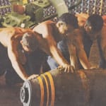 Four shirtless men roll a bomb-shaped cylinder.
