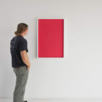 A man stands to the left and faces a red vertical rectangle in a white frame hanging on a white wall.