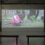 Still from video playing in gallery; a girl kneels alone in the grass.
